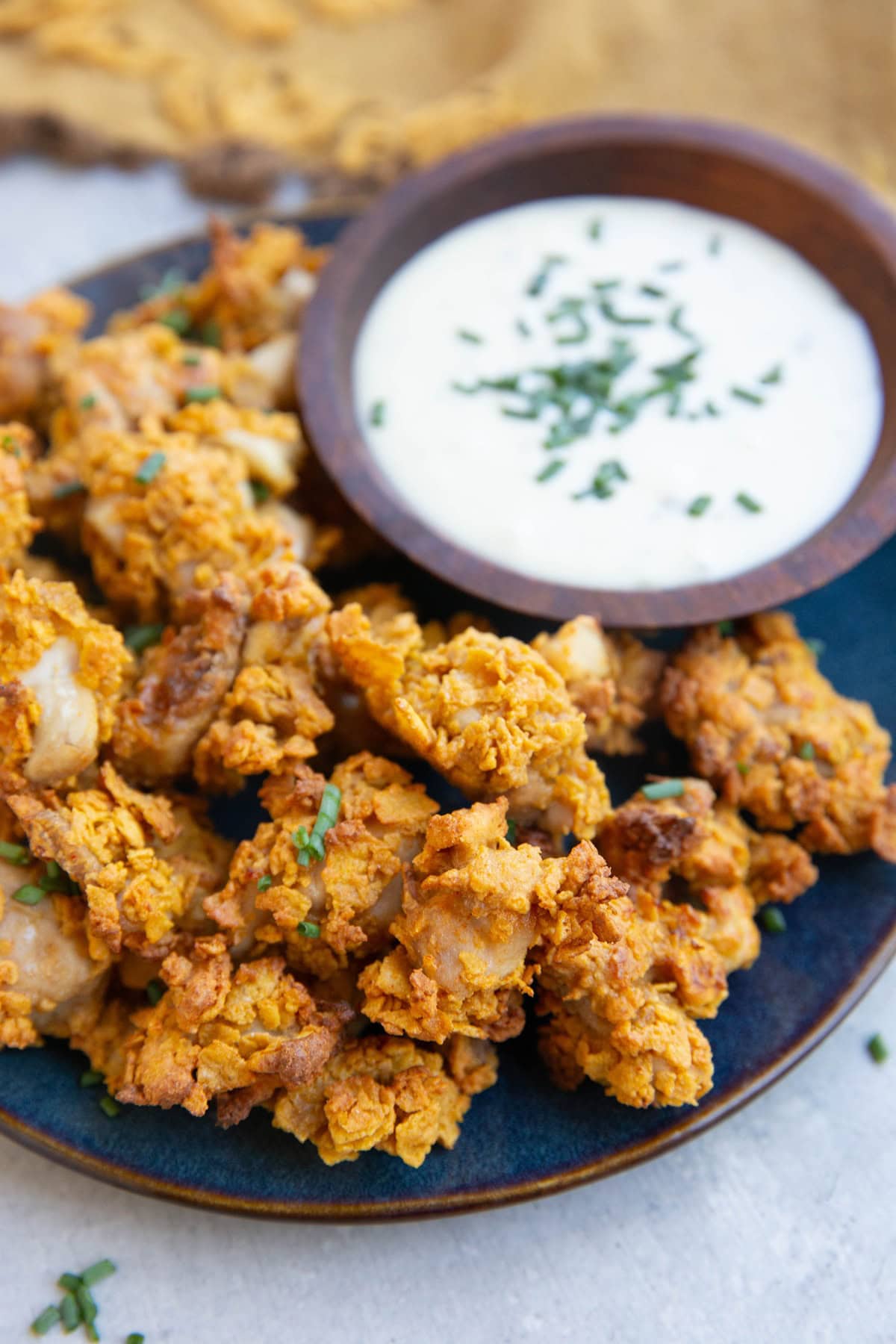 Blue plate full of popcorn chicken with a wooden bowl of ranch dressing and a golden napkin in the background.