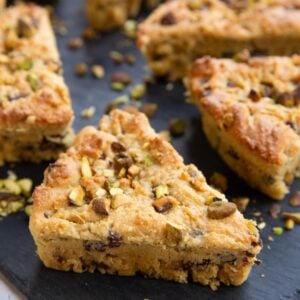Pistachio scones with pistachios sprinkled all around sitting on a black slate cutting board.