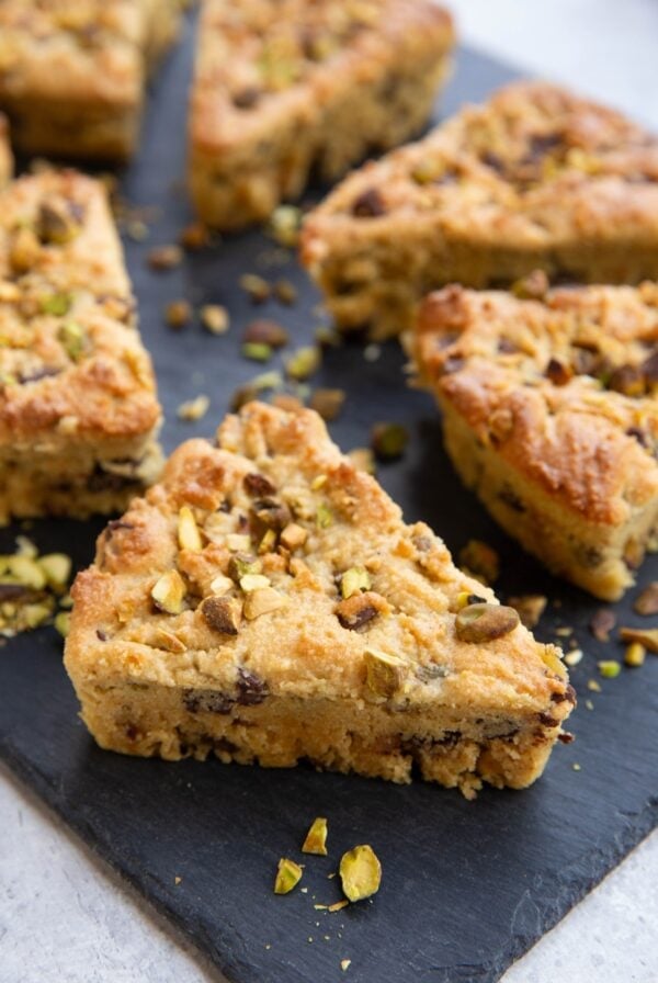 Pistachio scones with pistachios sprinkled all around sitting on a black slate cutting board.