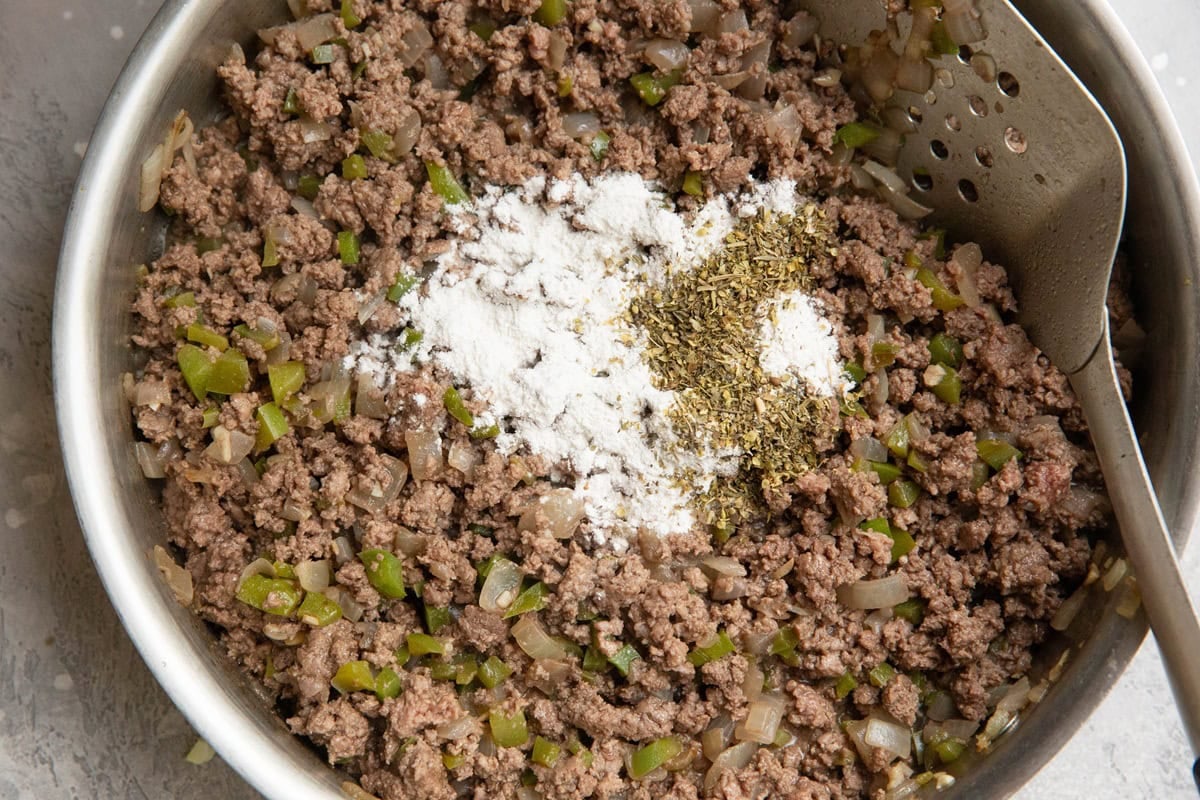 Skillet with cooked ground beef, onions and peppers with dried herbs and flour added into the skillet.