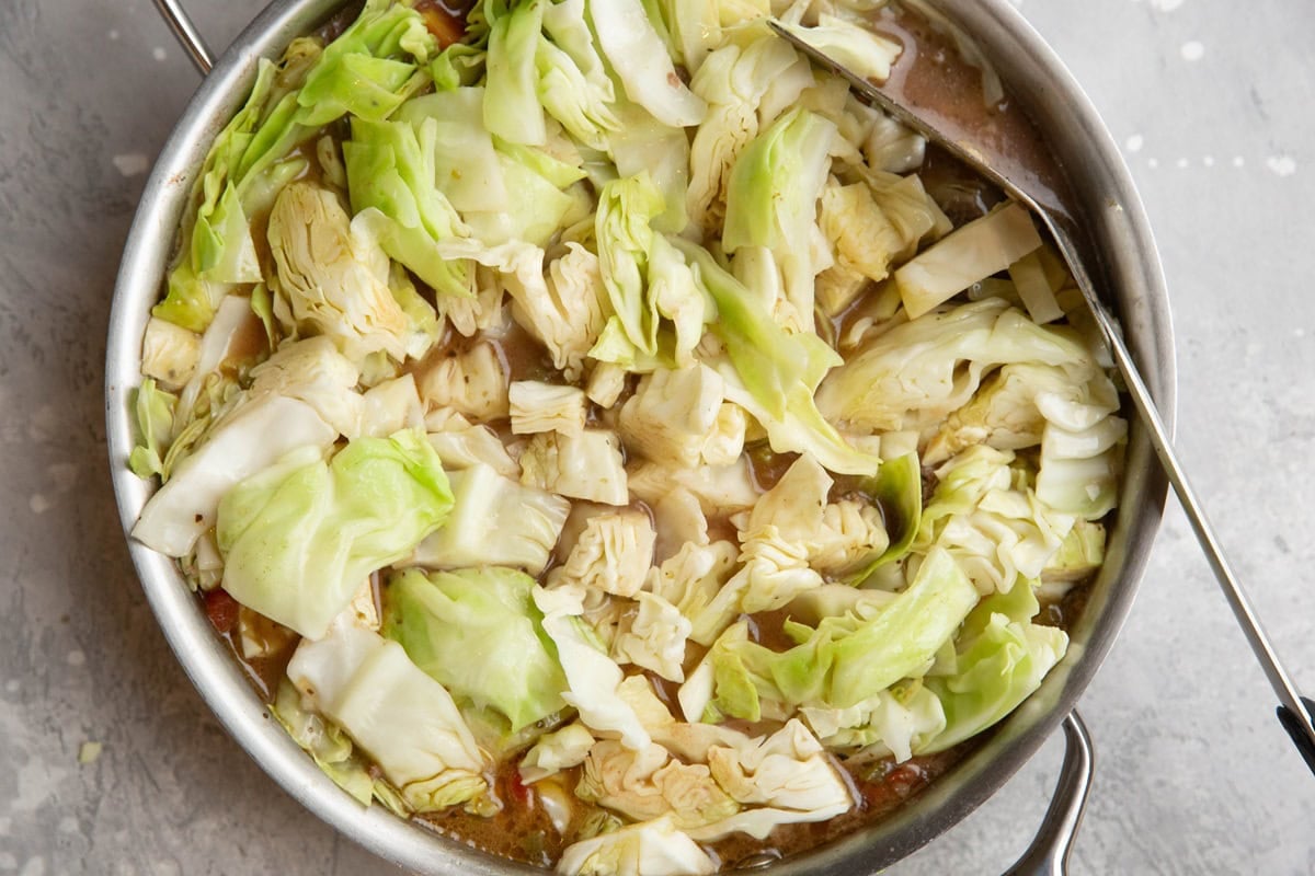Stainless steel skillet with chopped cabbage on top of liquid and meat, being cooked in.