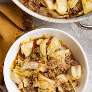 Skillet full of dirty cabbage and a white bowl full of dirty cabbage with a golden spoon and a golden napkin to the side.