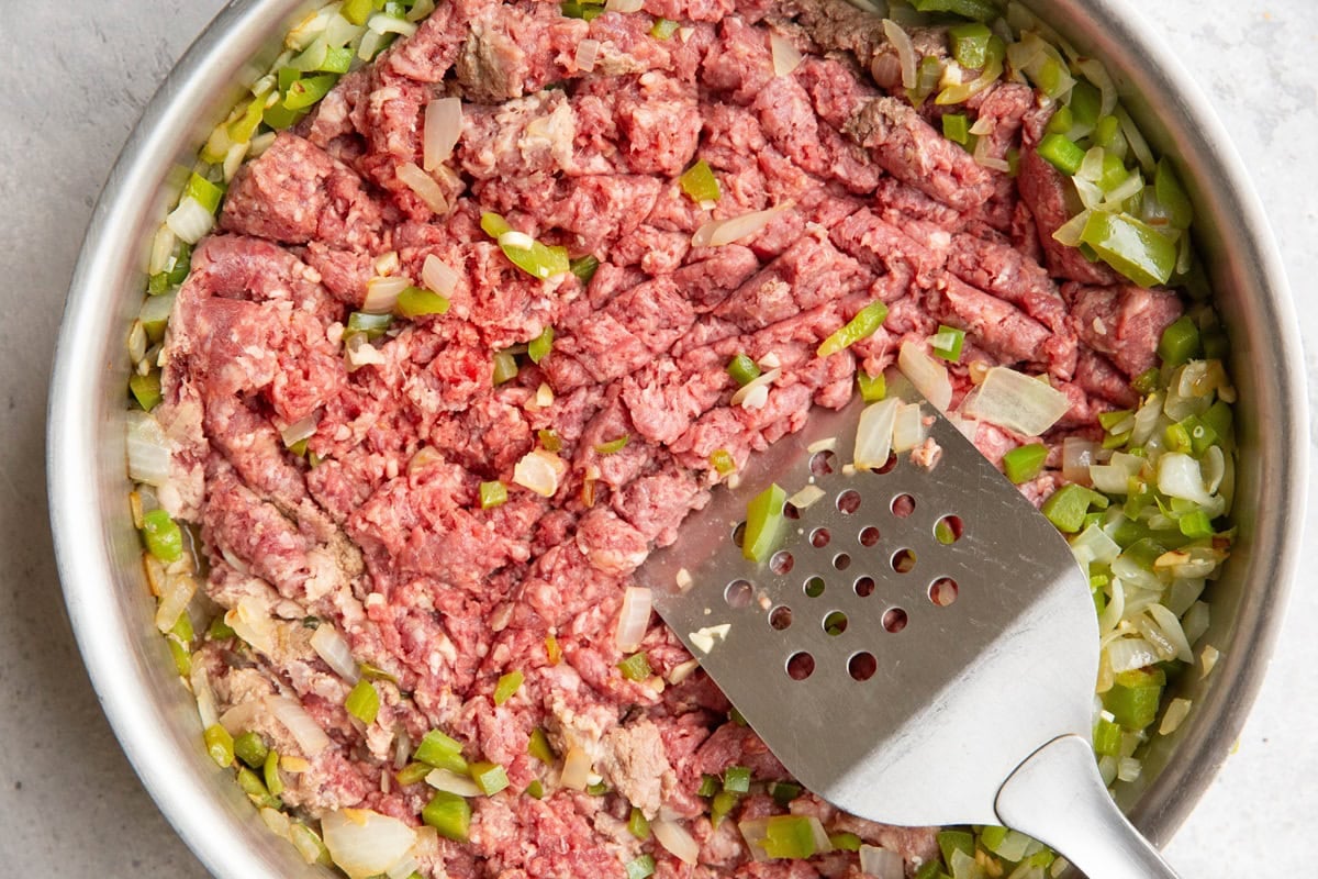 Stainless steel skillet with ground beef, onions, and peppers cooking inside and a spatula breaking up the meat into smaller chunks.