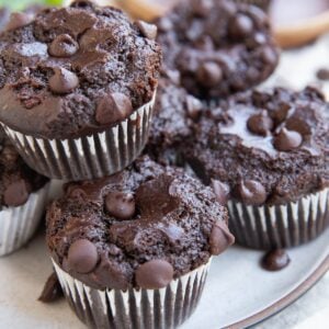 Plate with 7 chocolate muffins on it and a wooden plate with a single muffin in the background and a zucchini squash in the background.
