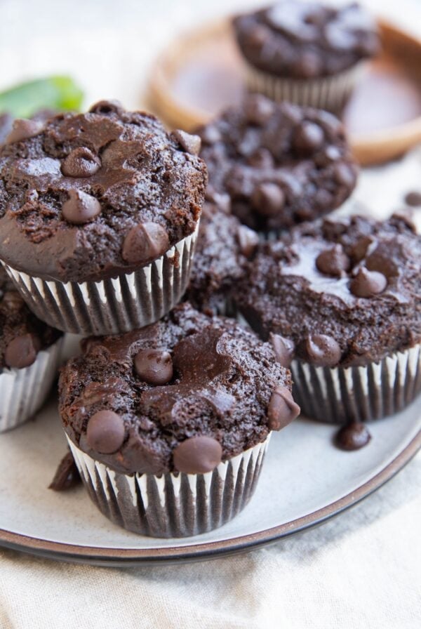 Plate with 7 chocolate muffins on it and a wooden plate with a single muffin in the background and a zucchini squash in the background.