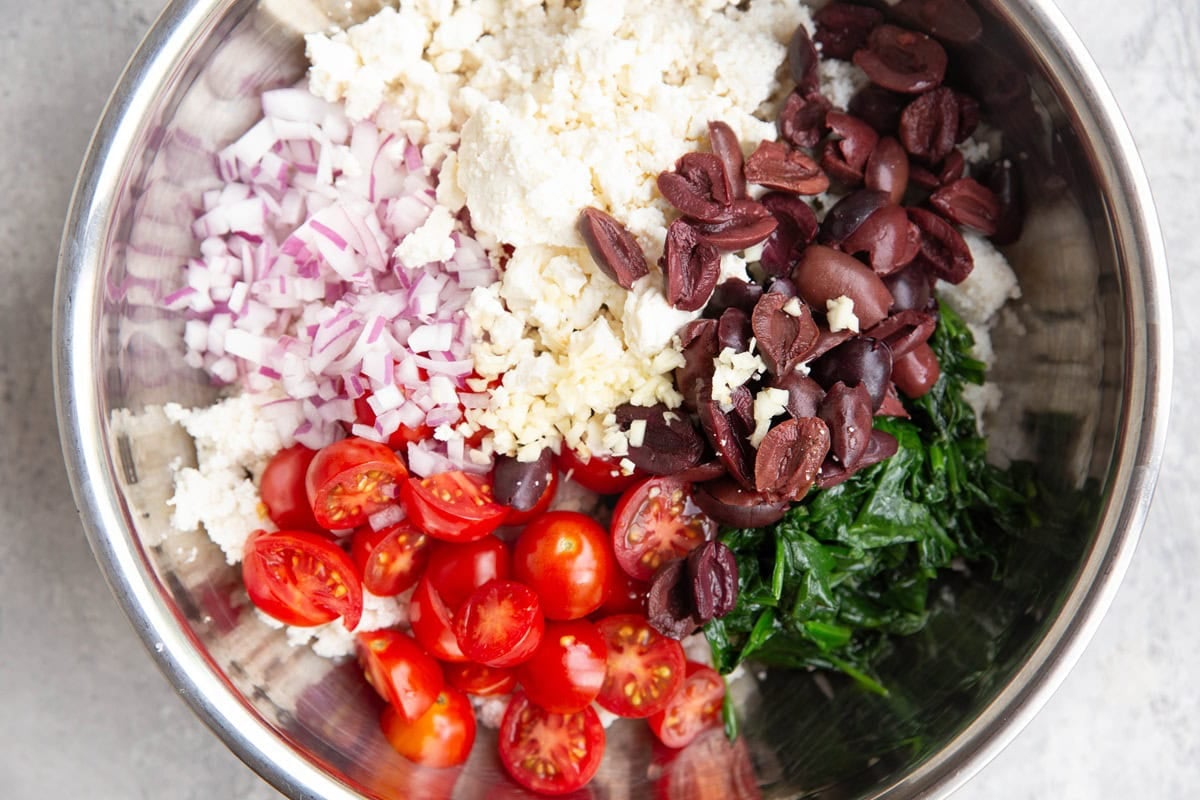 Stainless steel bowl full of cooked couscous, feta cheese, spinach, tomatoes, kalamata olives, and chopped red onion.