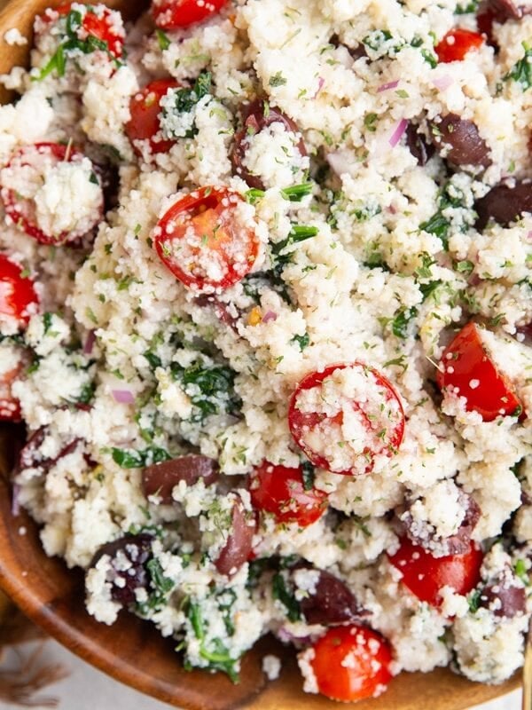 Wooden bowl full of Greek couscous salad with a golden napkin to the side and a golden spoon.