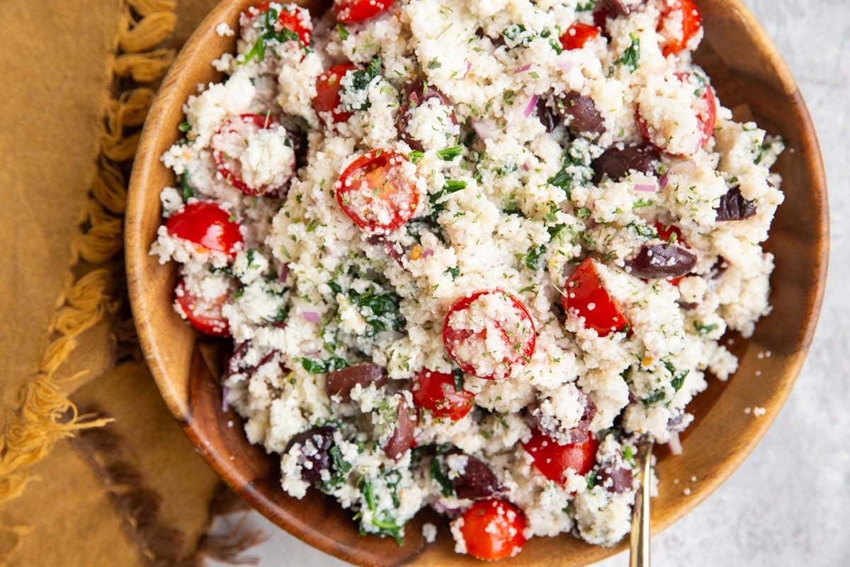 Wooden bowl full of Greek couscous salad with a golden napkin to the side and a golden spoon.