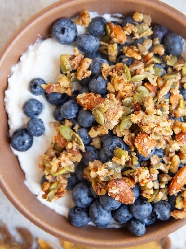 Bowl full of yogurt with blueberries and granola on top. A golden spoon sits inside of the bowl and a golden napkin to the side.