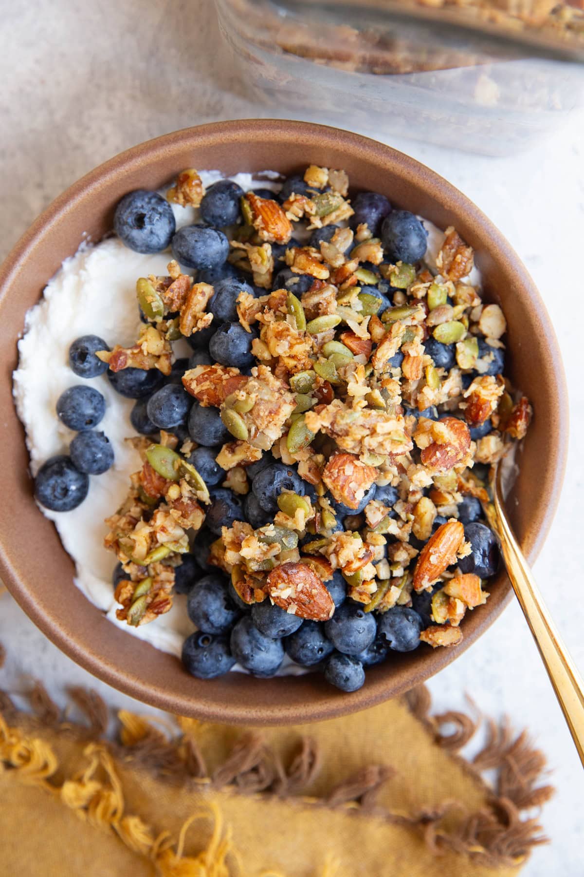 Brown bowl full of plain Greek yogurt, blueberries, and homemade granola with the jar of granola to the side.