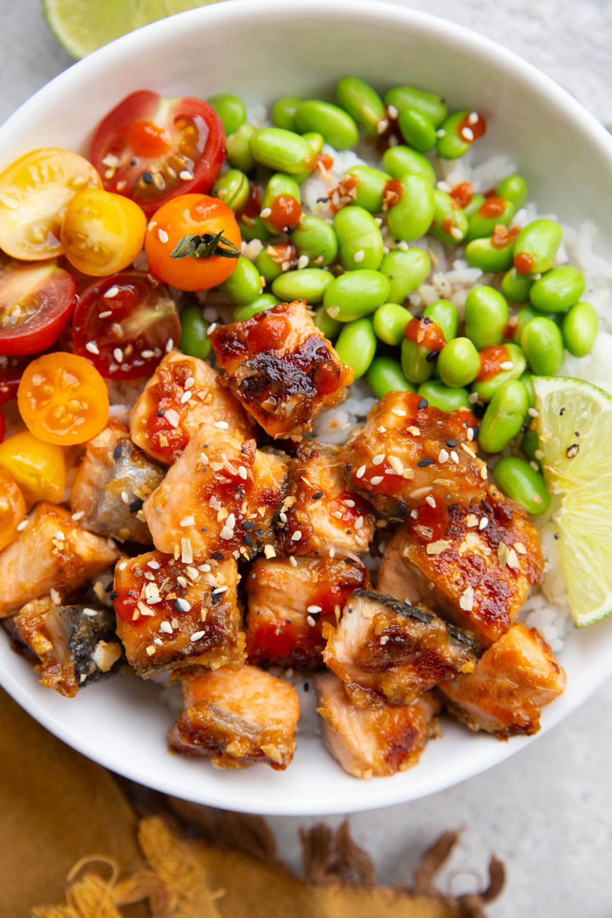 Miso salmon in a white bowl with edamame and chopped tomatoes. A golden napkin to the side. Ready to serve.