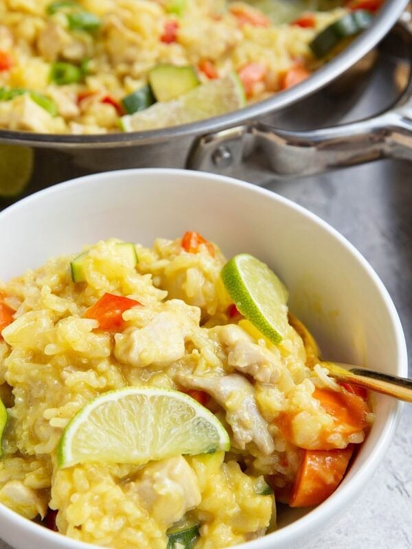 White bowl of chicken, rice, and vegetables in a coconut lime sauce with sliced limes on top. A stainless steel skillet is in the background with the rest of the meal inside and a fresh lime is cut in half to the side.