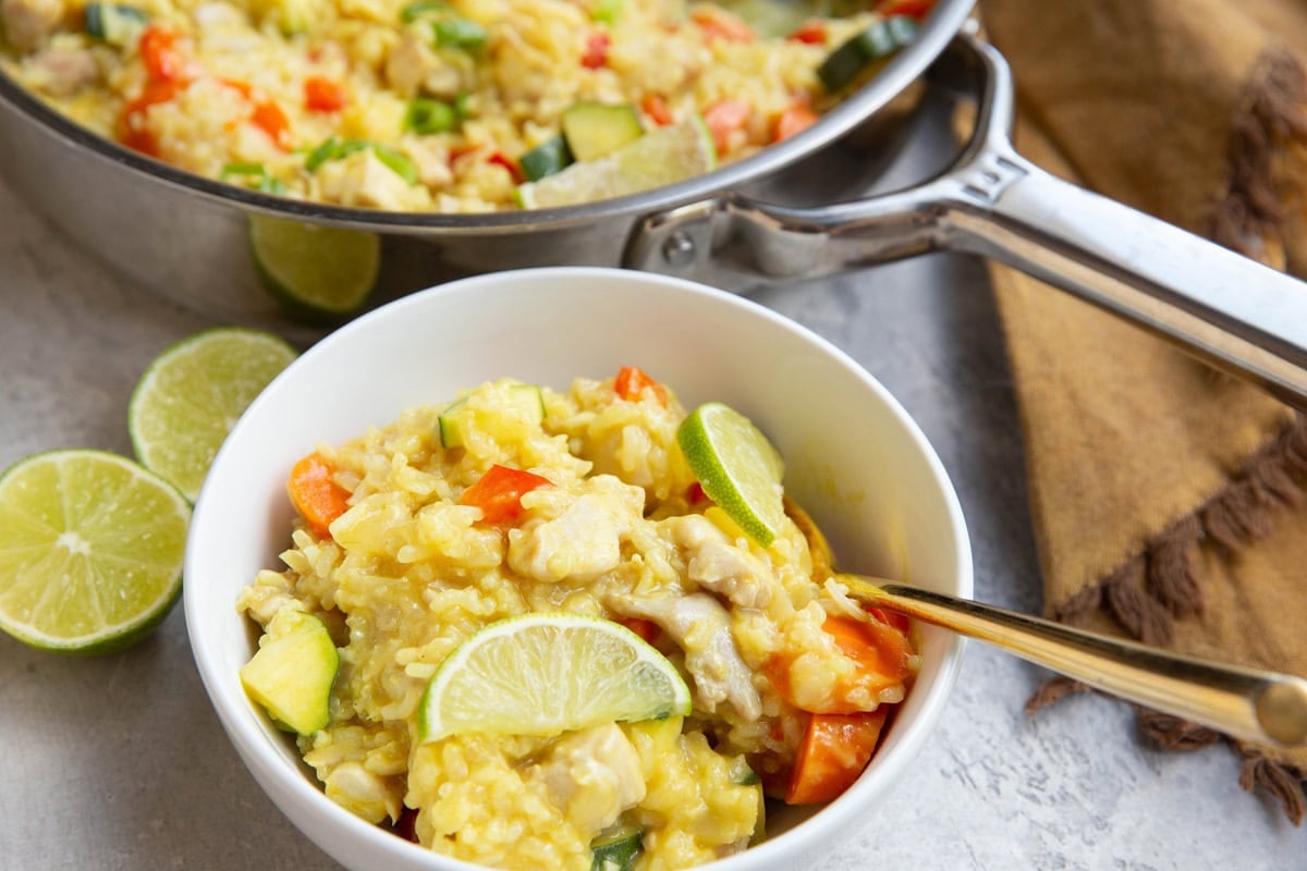 White bowl of chicken, rice, and vegetables in a coconut lime sauce with sliced limes on top. A stainless steel skillet is in the background with the rest of the meal inside and a fresh lime is cut in half to the side.