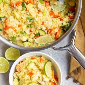 Stainless steel skillet with coconut lime chicken and rice with a wooden spoon to the side and a white bowl full of a portion of the meal. A golden napkin sits to the side, as does a fresh lime that has been cut in half.