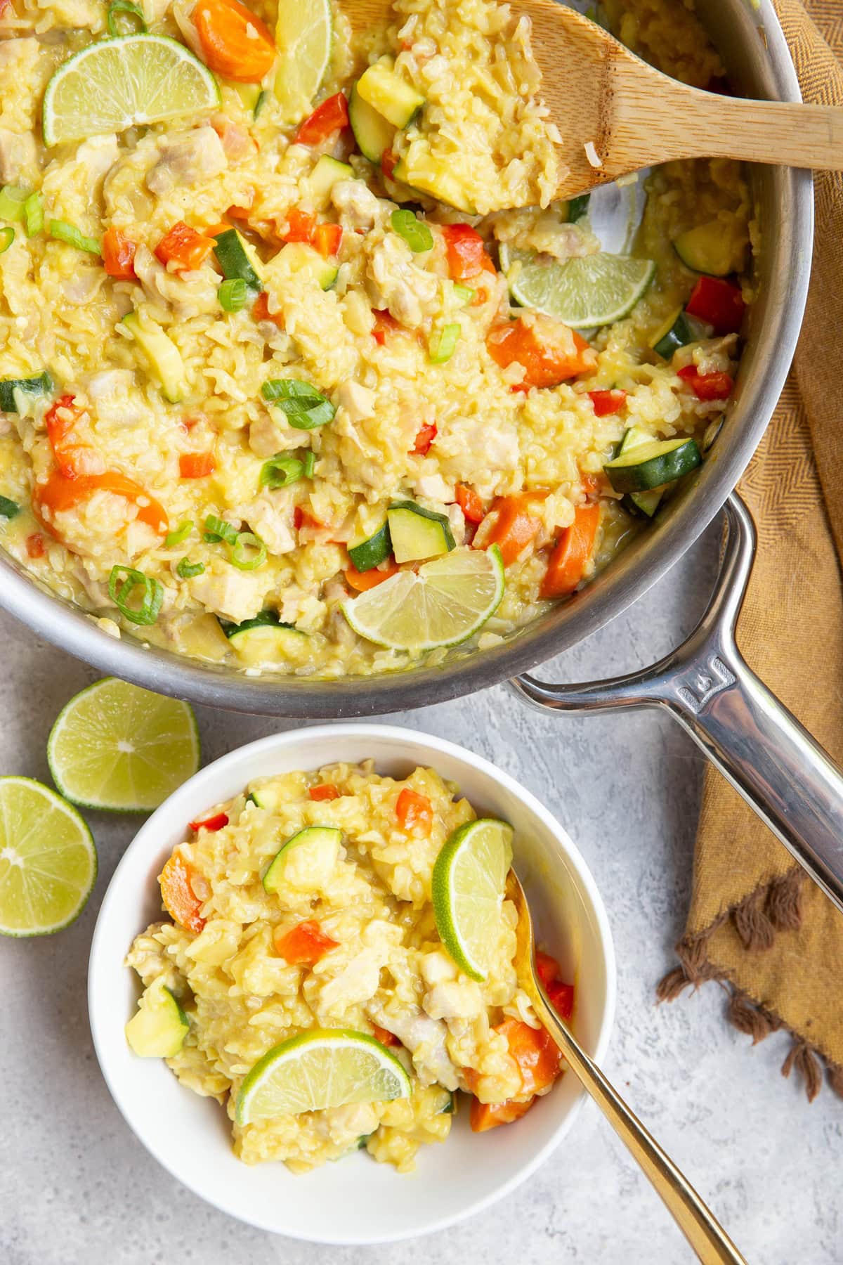 Stainless steel skillet with coconut lime chicken and rice with a wooden spoon to the side and a white bowl full of a portion of the meal. A golden napkin sits to the side, as does a fresh lime that has been cut in half.