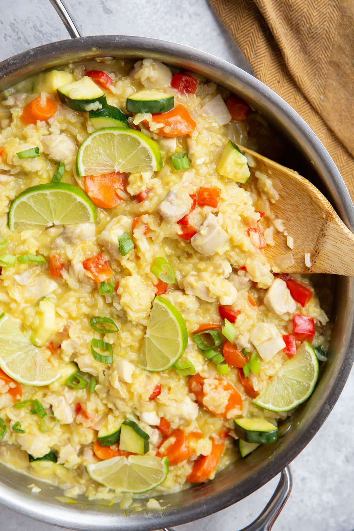 Stainless steel skillet full of coconut lime chicken with rice and vegetables with lime slices on top and chopped green onions. A wooden spoon sits inside the skillet for serving.