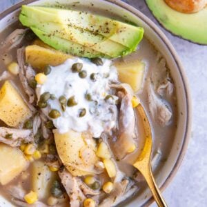 Bowl of chicken soup with chunks of potatoes, corn, capers, Greek yogurt on top and sliced avocado.