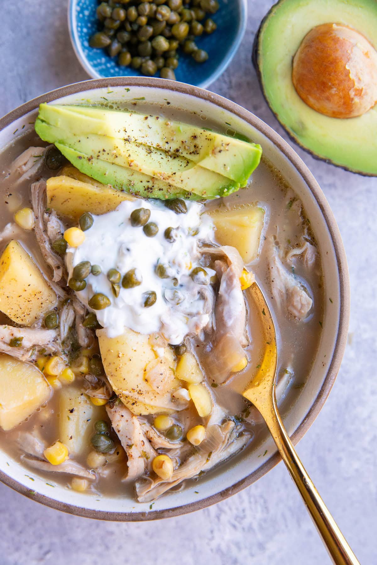 Bowl of chicken soup with chunks of potatoes, corn, capers, Greek yogurt on top and sliced avocado.