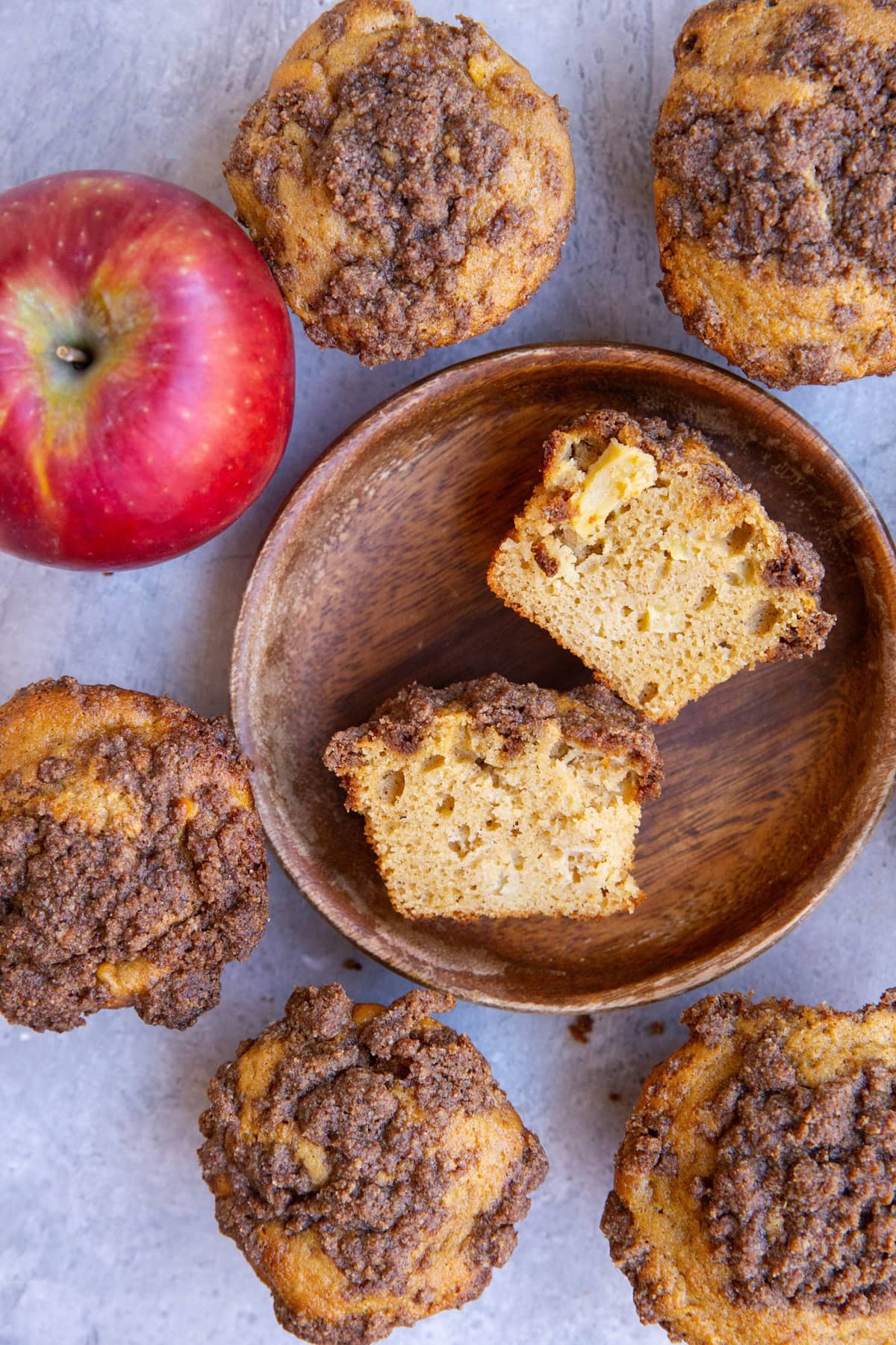 Wooden plate with an apple muffin cut in half and more apple crumb muffins around the plate and a fresh apple to the side.