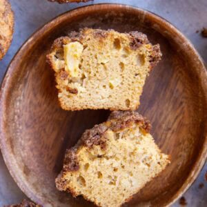 Apple muffins sliced on a wooden plate in half to expose the inside of the muffin. More muffins sitting around the wooden plate.