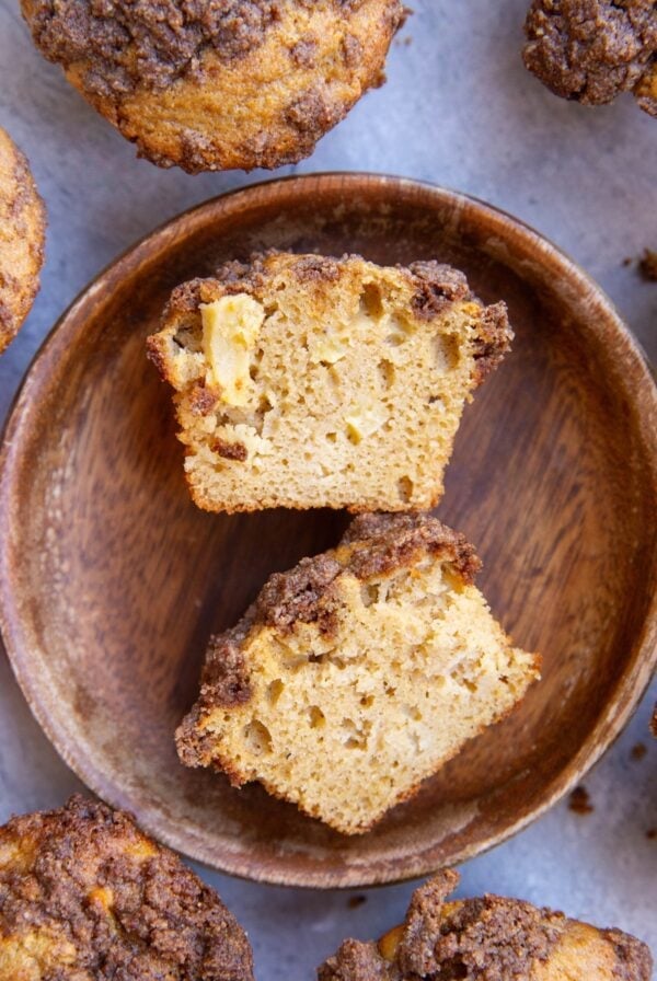 Apple muffins sliced on a wooden plate in half to expose the inside of the muffin. More muffins sitting around the wooden plate.