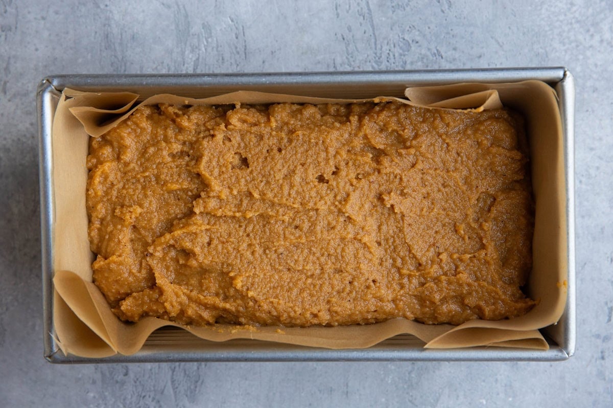 Loaf pan lined in parchment paper with pumpkin bread batter spread inside.