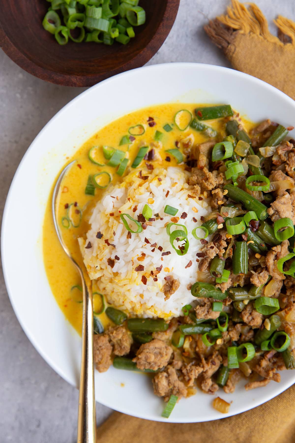 White bowl full of rice, ground turkey, green beans, and a yellow coconut curry broth, sprinkled with green onions and red pepper flakes, and a golden spoon to the side.