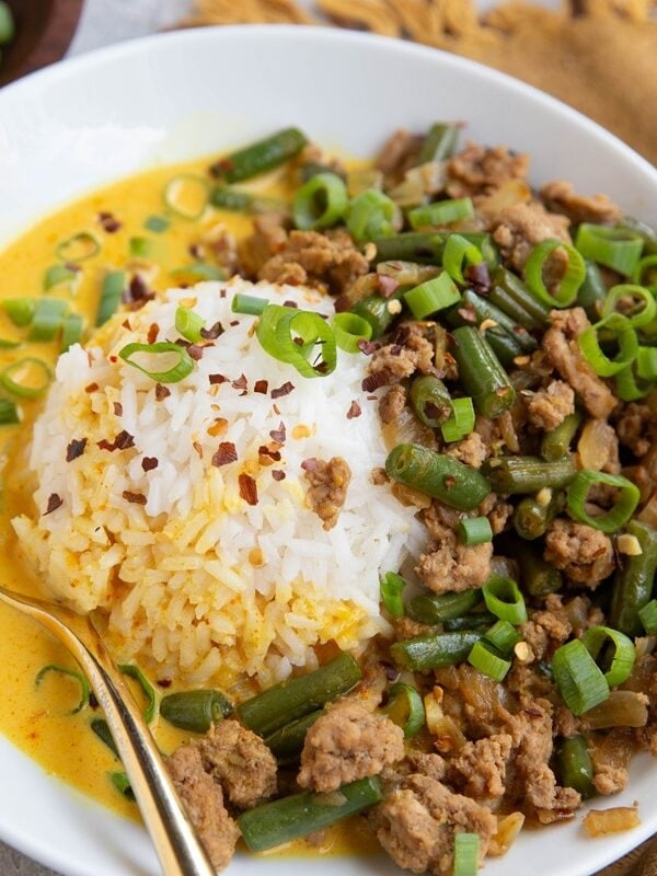 Ground turkey and green beans on one side of a white bowl with a scoop of white rice in the middle and a yellow broth surrounding everything. A golden napkin in the background and a bowl of chopped green onions.