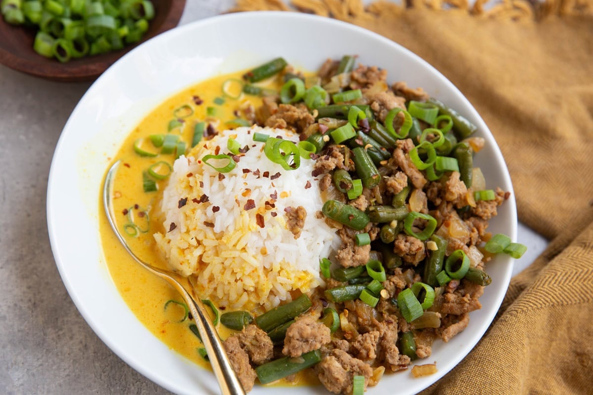 Ground turkey and green beans on one side of a white bowl with a scoop of white rice in the middle and a yellow broth surrounding everything. A golden napkin in the background and a bowl of chopped green onions.
