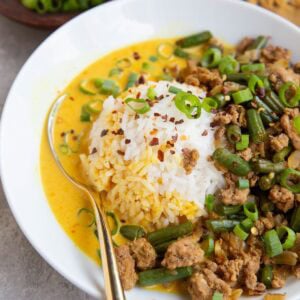 Ground turkey, green beans, a scoop of white rice and a yellow broth sit in a white bowl that is garnished with chopped green onions and red pepper flakes.