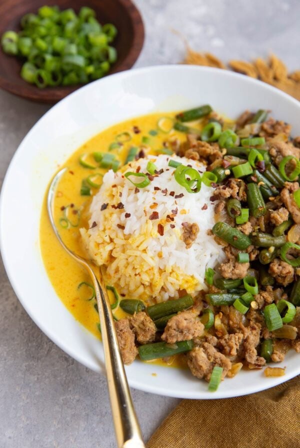 Ground turkey, green beans, a scoop of white rice and a yellow broth sit in a white bowl that is garnished with chopped green onions and red pepper flakes.