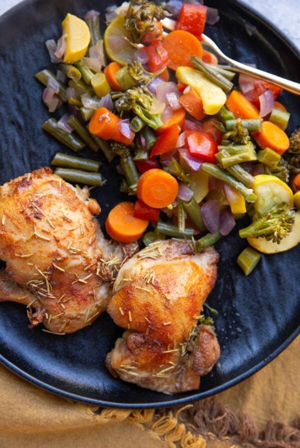 Cooked chicken thighs and roasted vegetables on a black plate with a gold fork.
