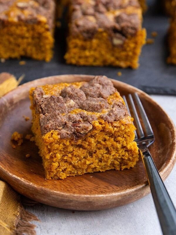 Slice of pumpkin cake on a wooden plate with a black slate cutting board with slices of cake on top in the background and a golden napkin to the side.
