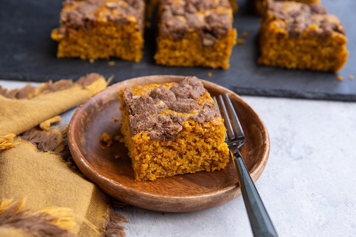 Slice of pumpkin cake on a wooden plate with a black slate cutting board with slices of cake on top in the background and a golden napkin to the side.