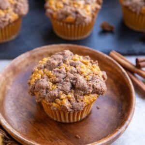 Pumpkin muffin on a wooden plate with streusel topping and the rest of the muffins on a black cutting board in the background.