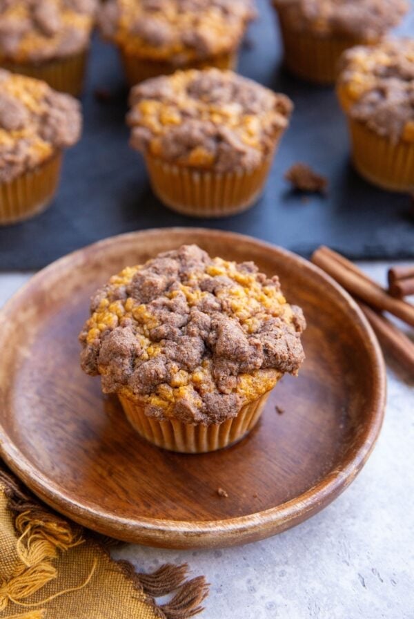Pumpkin muffin on a wooden plate with streusel topping and the rest of the muffins on a black cutting board in the background.