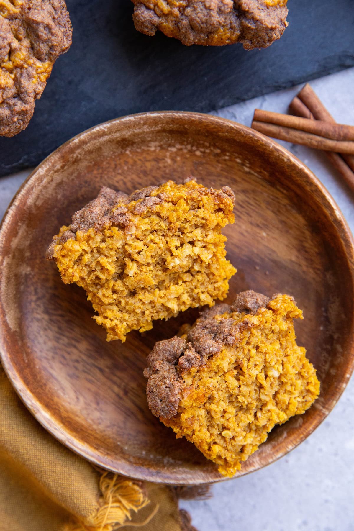 Pumpkin muffin cut in half on a wooden plate with the rest of the muffins and some cinnamon sticks to the side.
