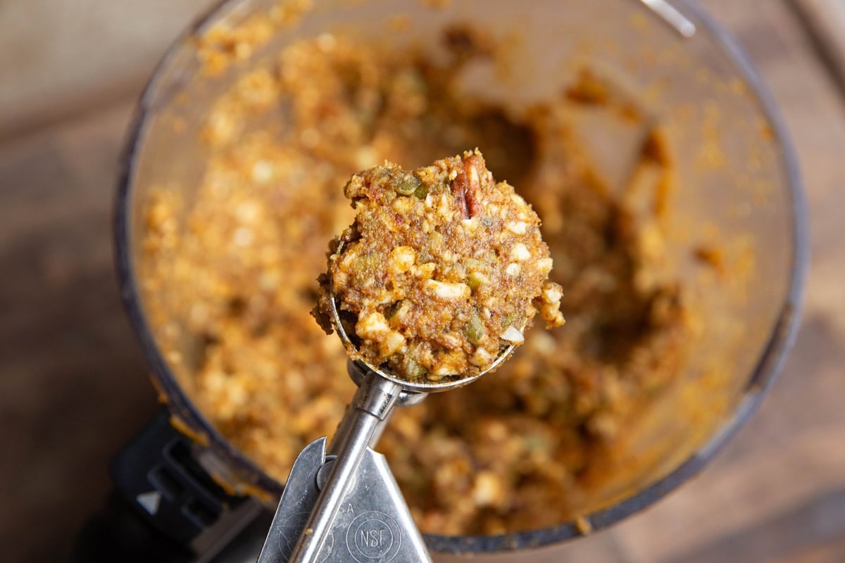 Cookie scoop with some pumpkin protein ball mixture inside hovering above the food processor full of the dough.