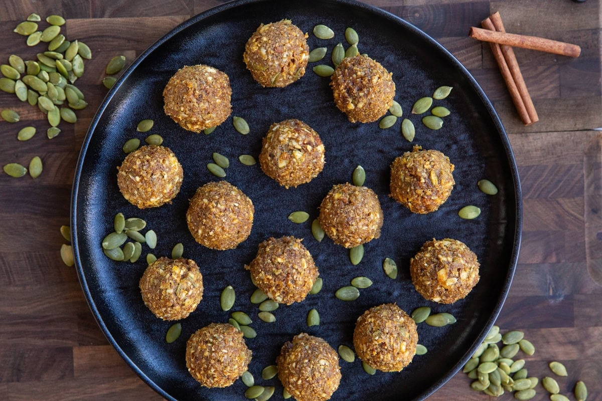 Black plate of pumpkin protein balls with pumpkin seeds sprinkled around on a wooden cutting board with cinnamon sticks.