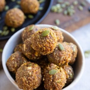 White bowl full of pumpkin protein balls with a black plate full of pumpkin protein balls in the background.
