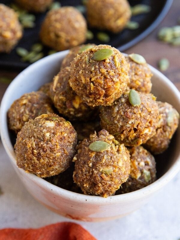 White bowl of pumpkin protein balls with pumpkin seeds scattered around and the plate of protein balls