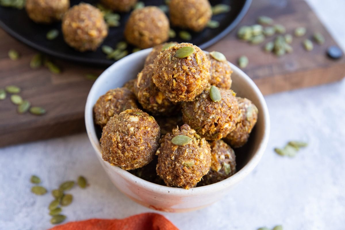 White bowl of pumpkin protein balls with pumpkin seeds scattered around and the plate of protein balls