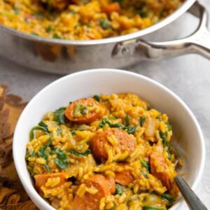White bowl with a serving of sausage, rice, and spinach with pumpkin and a fork to the side. A large skillet with the rest of the skillet in the background.
