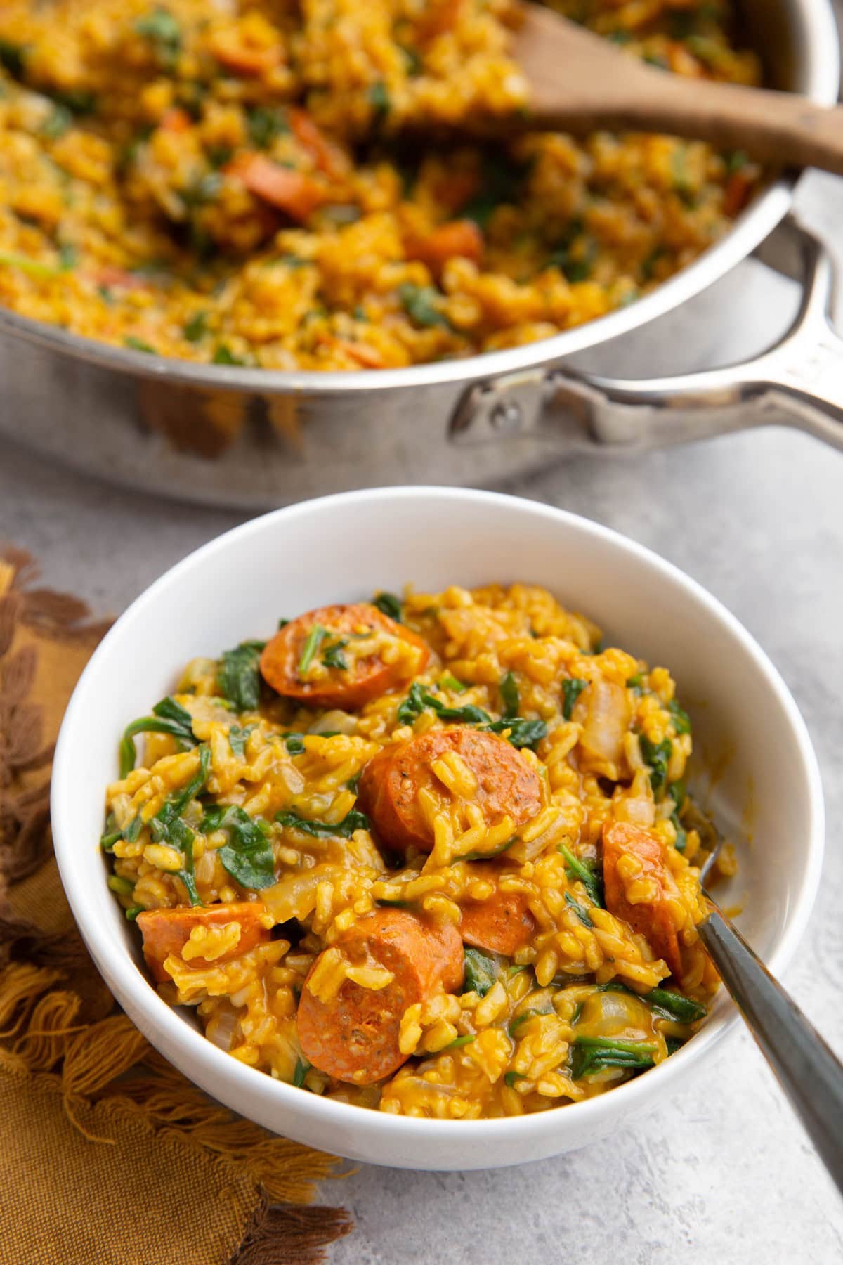 White bowl with a serving of sausage, rice, and spinach with pumpkin and a fork to the side. A large skillet with the rest of the skillet in the background.
