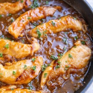 Skillet full of 7 cooked chicken tenderloins in a dark brown caramelized onion gravy.