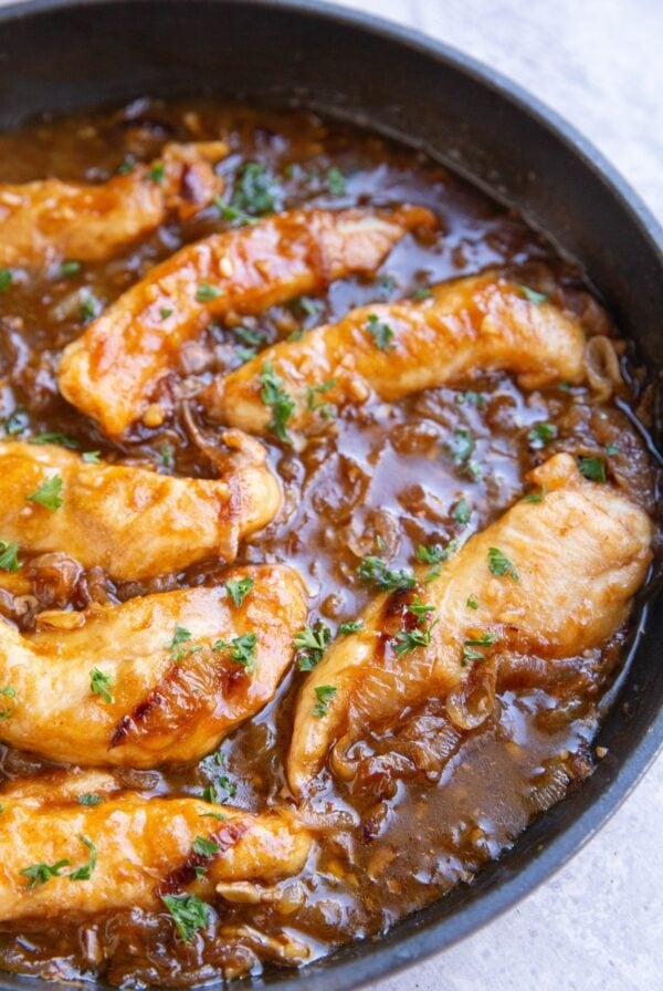 Skillet full of 7 cooked chicken tenderloins in a dark brown caramelized onion gravy.