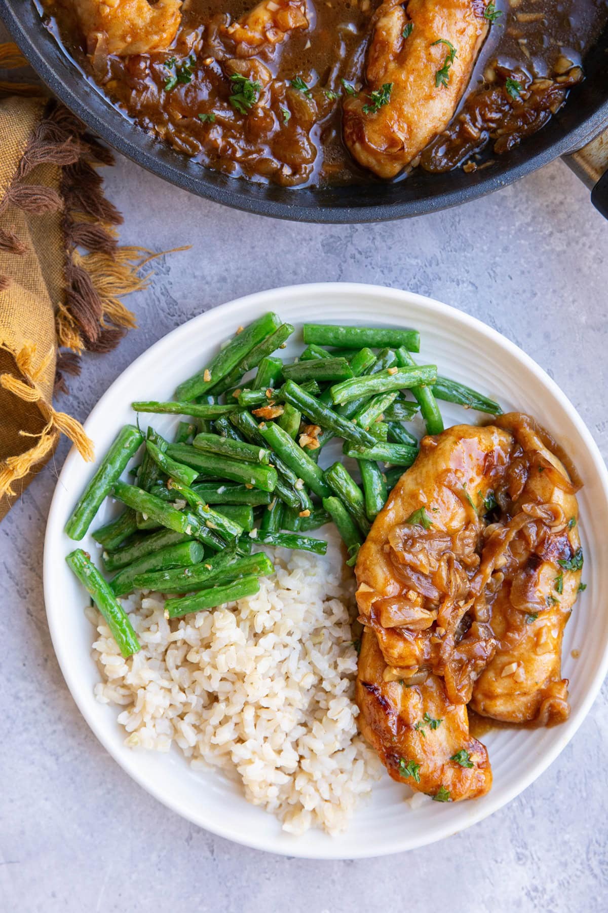 Plate of tender chicken with caramelized onion sauce on top paired with brown rice and green beans and the skillet with the rest of the saucy chicken to the side.