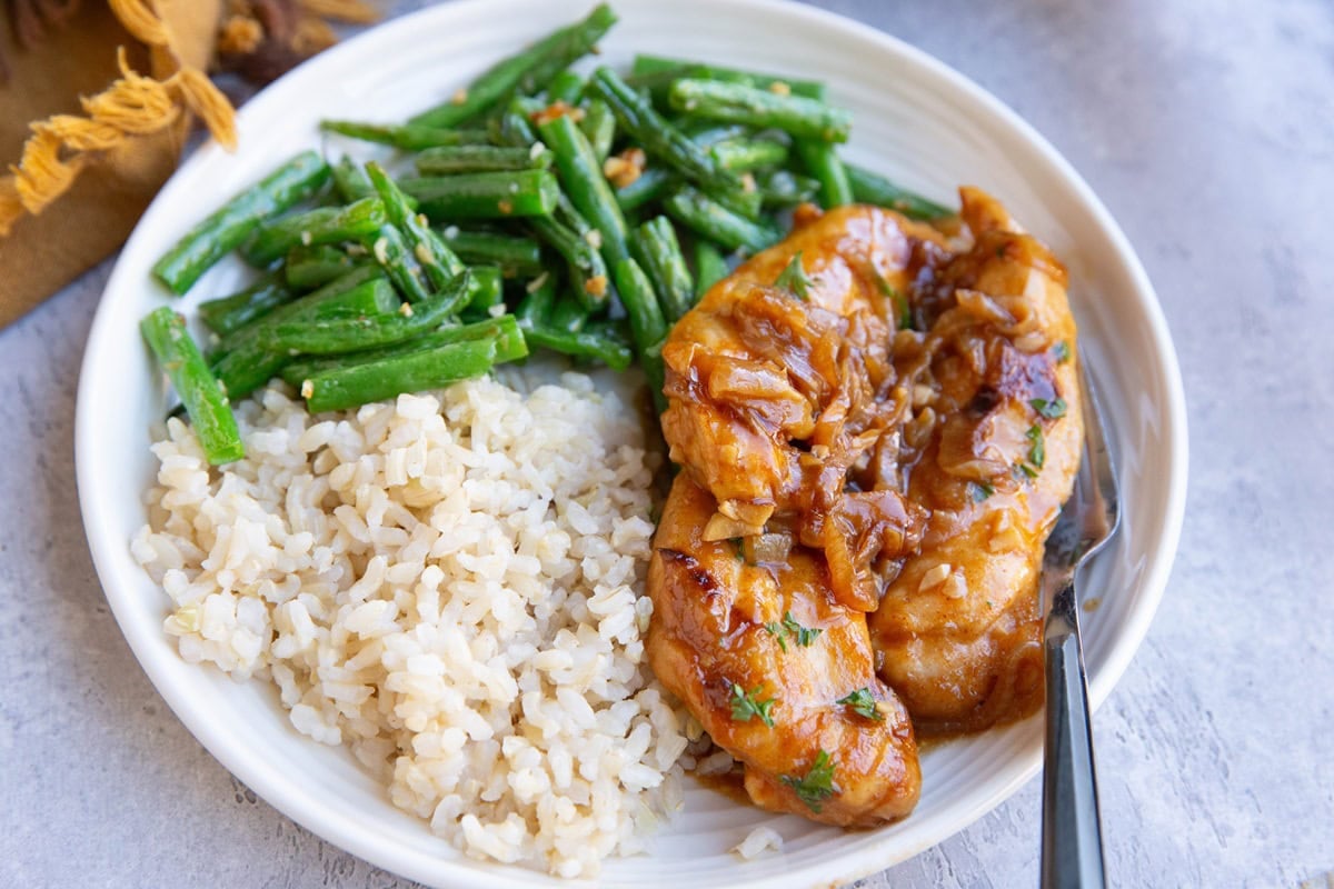 White plate of tender smothered chicken in caramelized onion sauce with brown rice and green beans