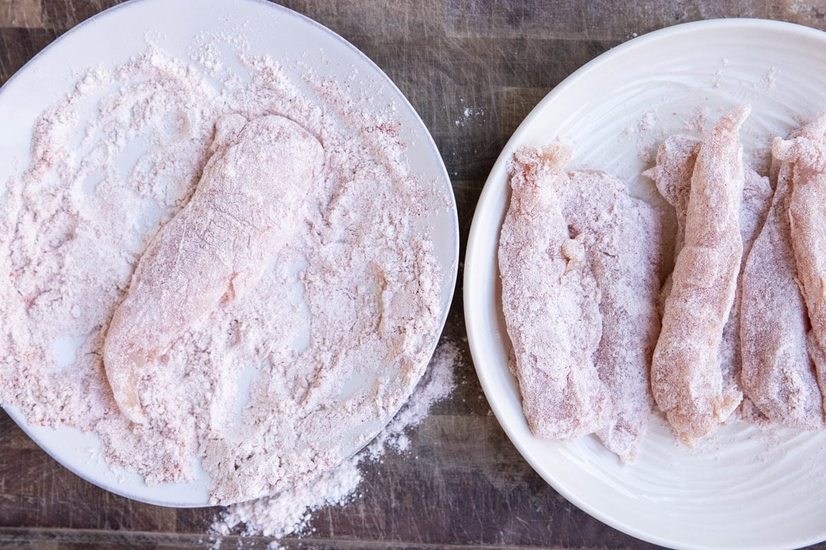 Plate of many raw chicken tenders dredged in flour on the right and a plate with a raw chicken tender being dipped in a flour mixture to make breaded chicken.