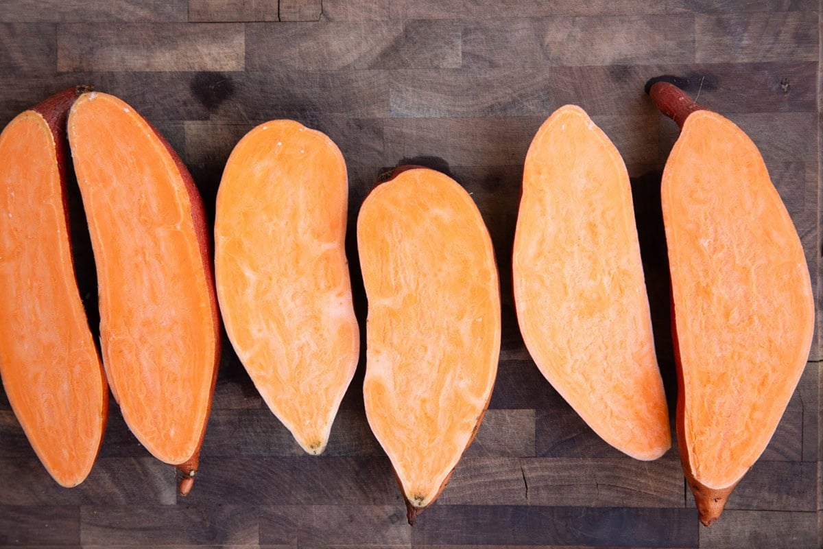 Three sweet potatoes cut in half lengthwise on a wooden cutting board.